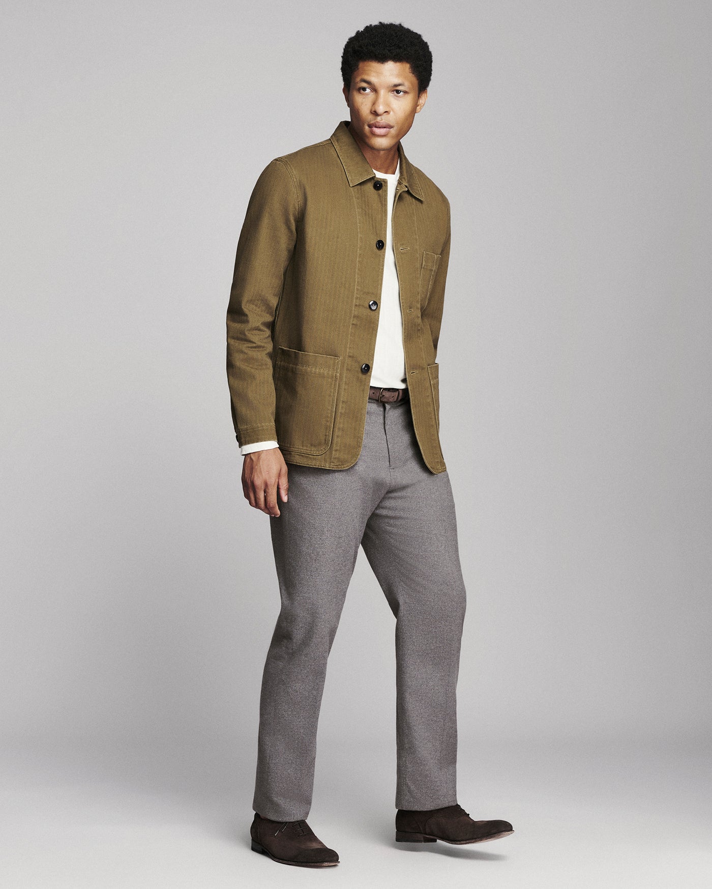 A man modeling a brown corduroy utility jacket over a white shirt, paired with gray trousers and dark brown shoes, against a light gray studio background.
