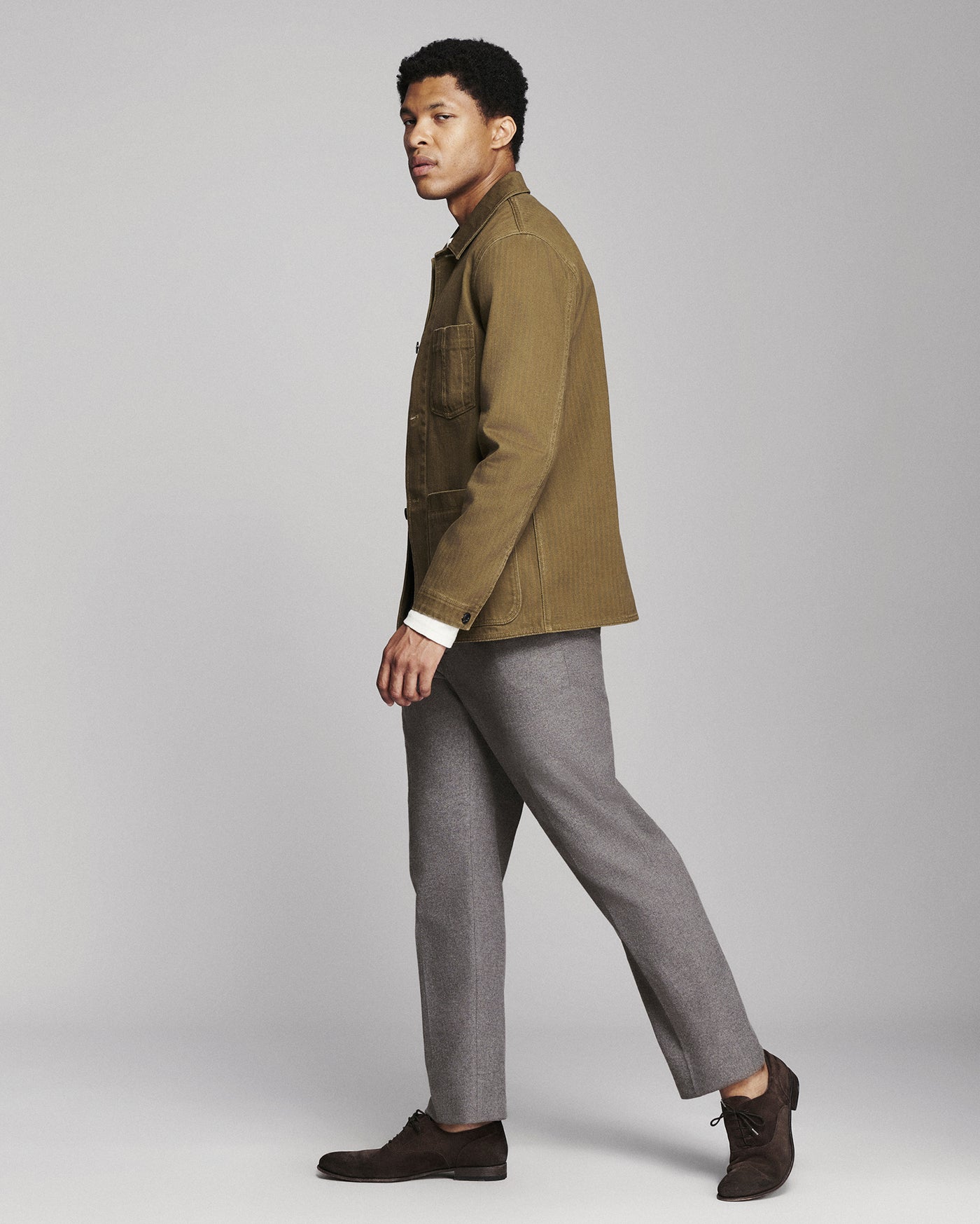 A man in side profile wearing a brown corduroy jacket, gray trousers, and dark brown suede shoes, standing against a light gray studio background.
