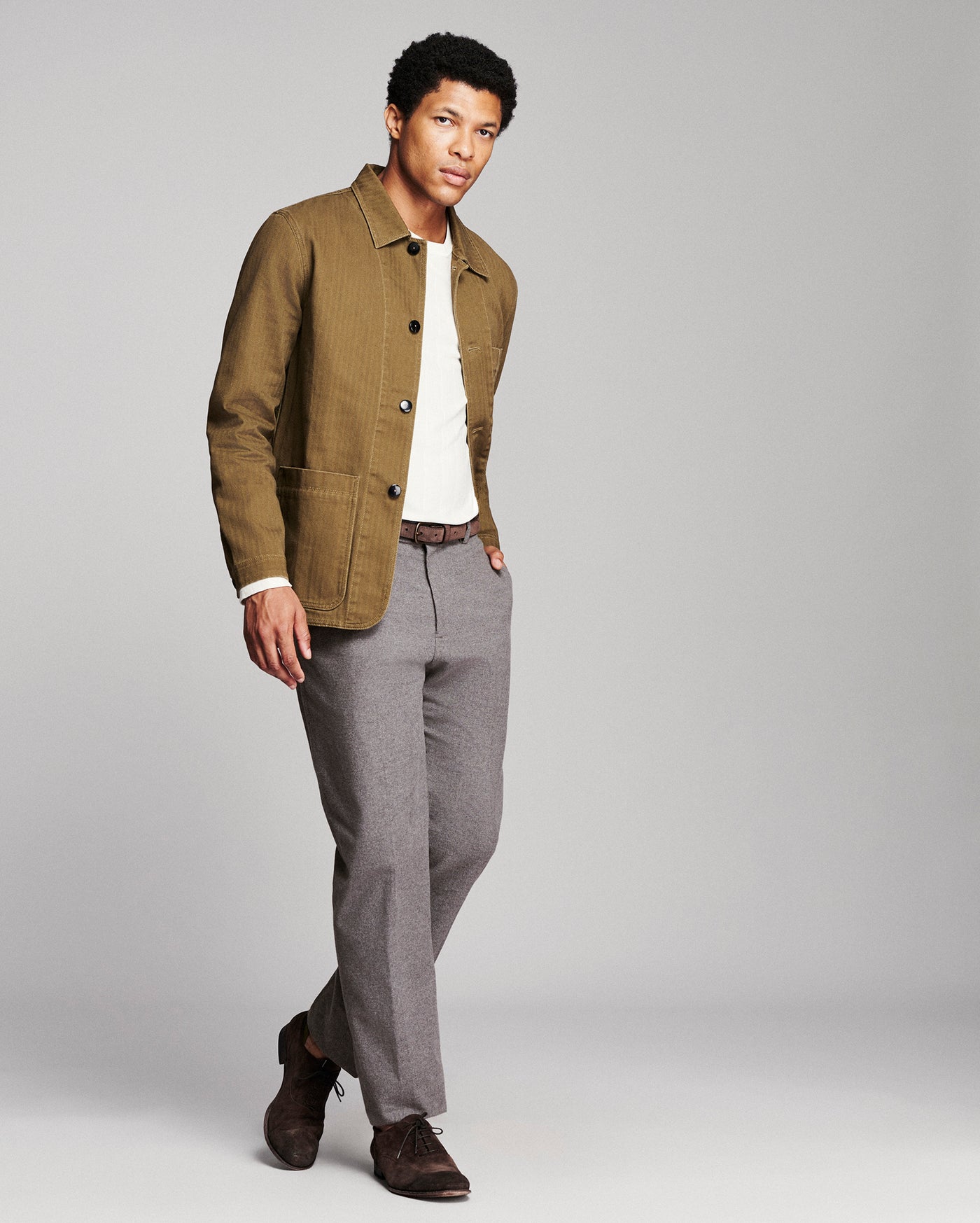 A man modeling a brown corduroy jacket over a white t-shirt, paired with gray trousers and brown suede shoes, against a solid gray studio background.
