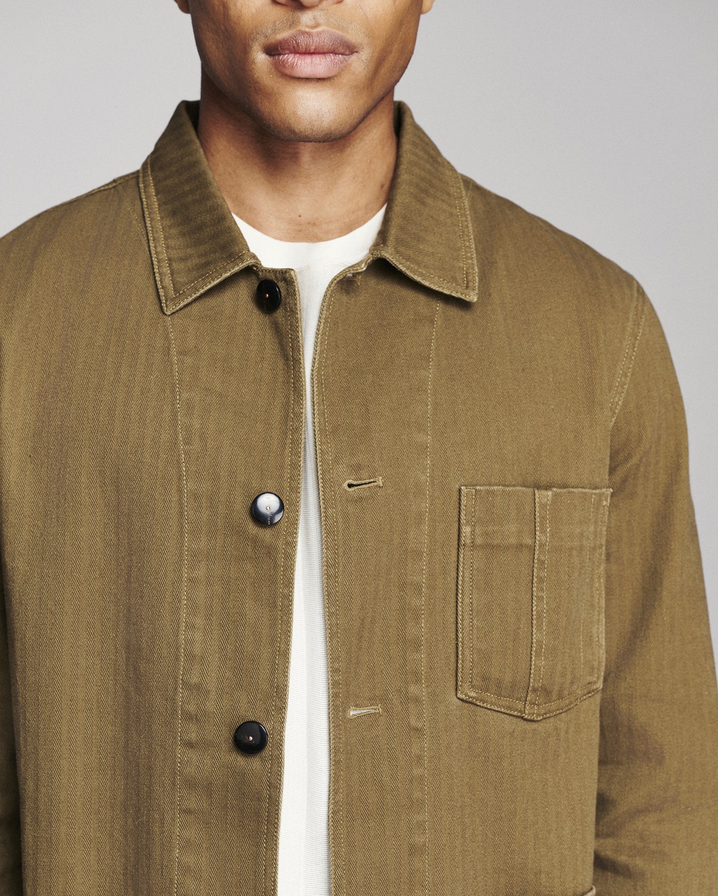 A close-up of a man wearing a brown corduroy utility jacket over a white t-shirt, showing the collar, front buttons, and chest pocket.