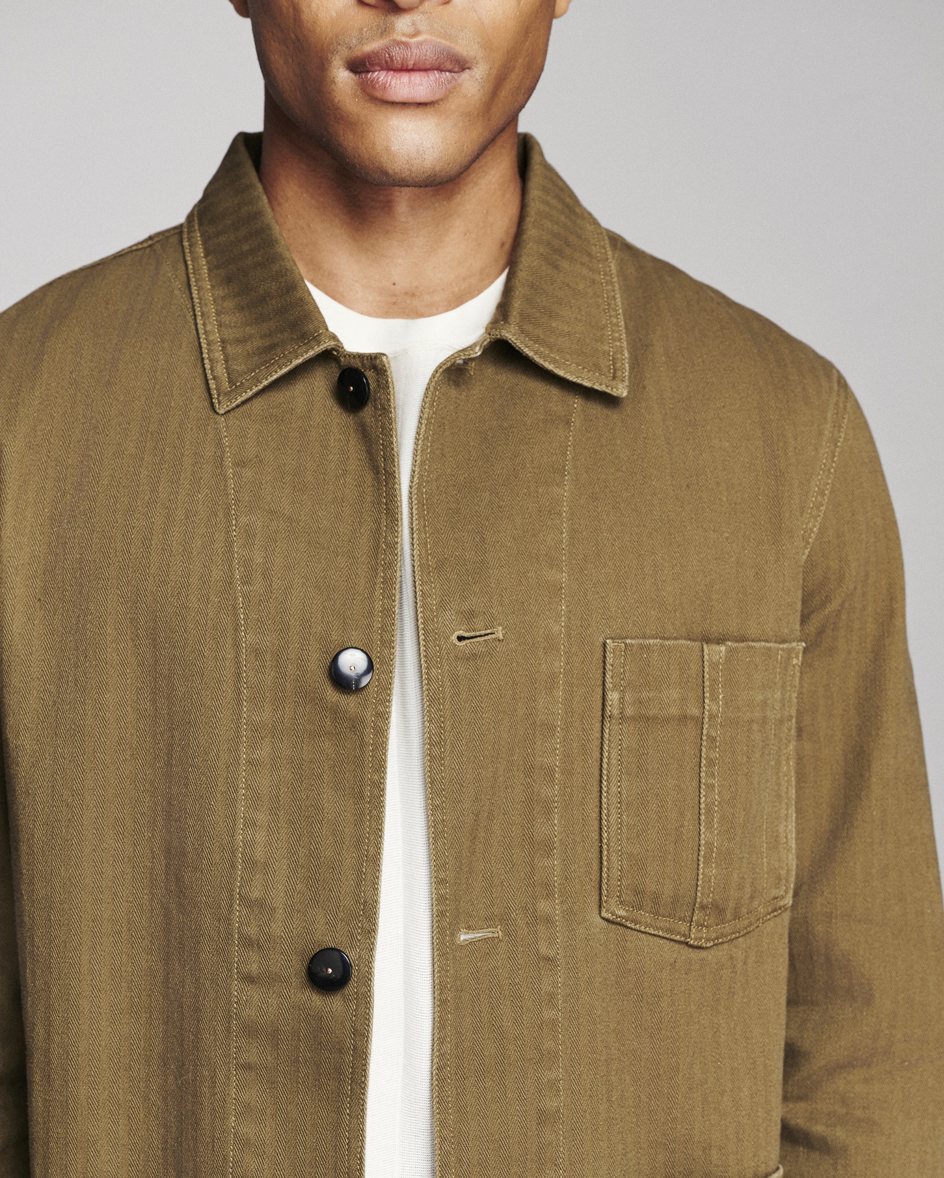 A close-up of a man wearing a brown corduroy utility jacket over a white t-shirt, showing the collar, front buttons, and chest pocket.