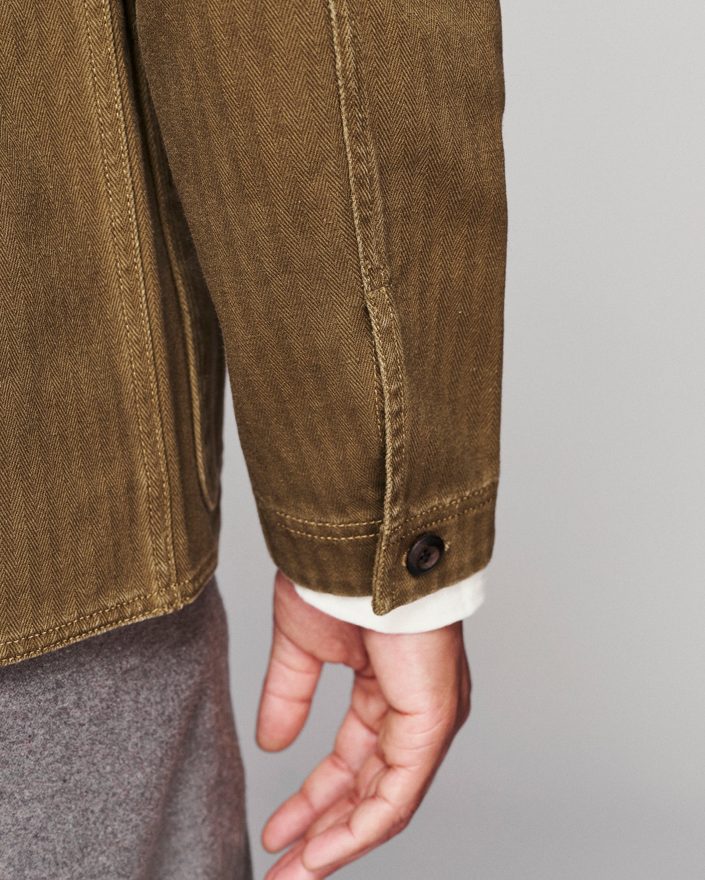 Close-up of a person wearing an olive brown canvas utility jacket, showing the sleeve, button cuff, and hand against a light gray background.