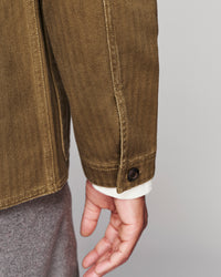 Close-up of a person wearing an olive brown canvas utility jacket, showing the sleeve, button cuff, and hand against a light gray background.