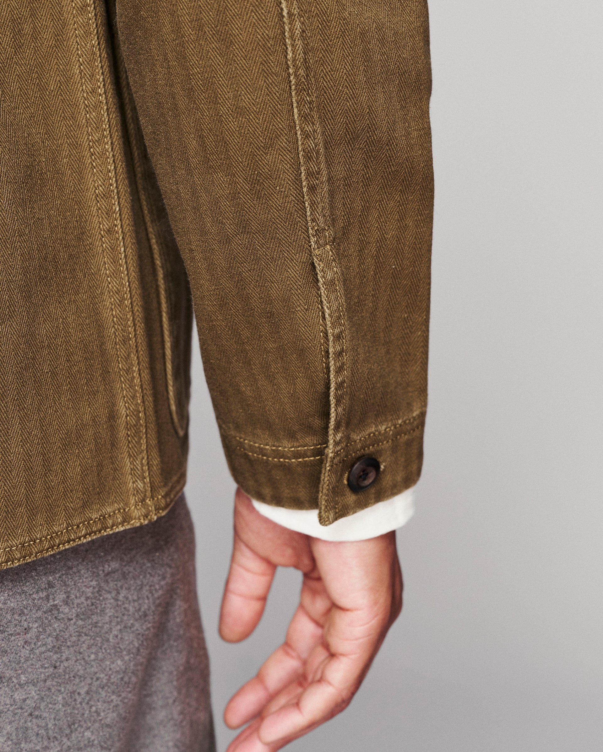 Close-up of a person wearing an olive brown canvas utility jacket, showing the sleeve, button cuff, and hand against a light gray background.