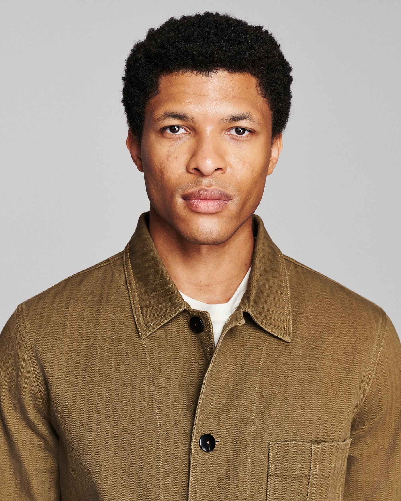 A man wearing a brown corduroy button-up shirt over a white undershirt, photographed against a solid gray background.