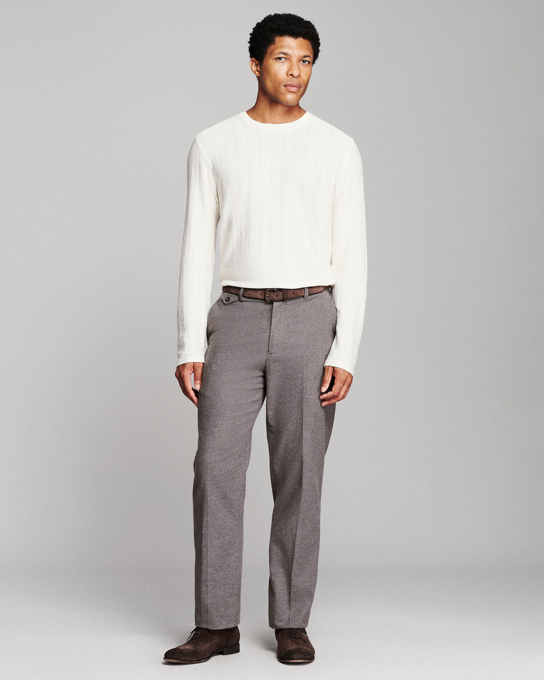 A man modeling a white ribbed long-sleeve knit sweater and gray pleated trousers with a brown belt and dark brown shoes, standing against a light gray studio background.