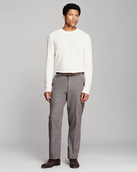 A man modeling a white ribbed long-sleeve knit sweater and gray pleated trousers with a brown belt and dark brown shoes, standing against a light gray studio background.