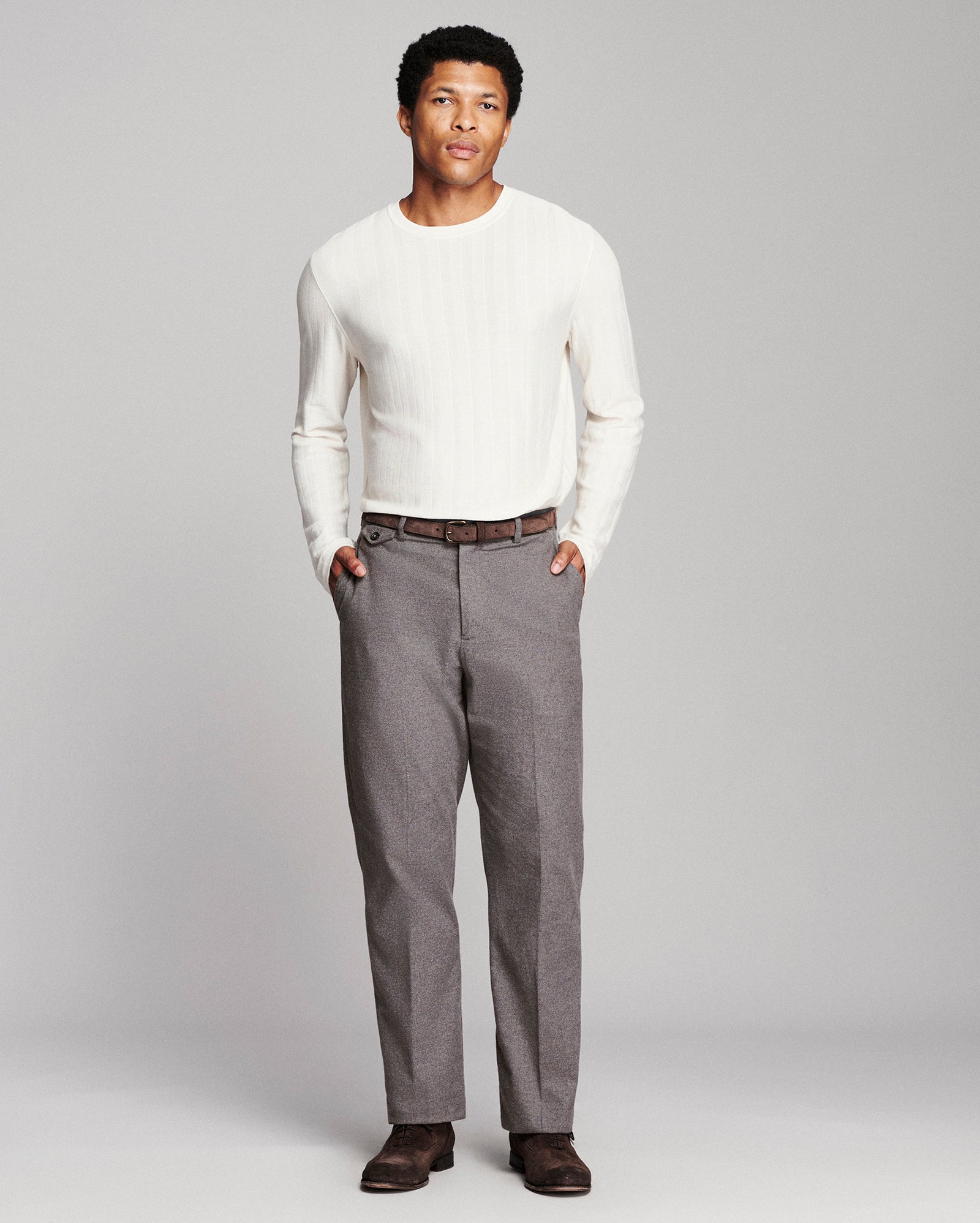 A man modeling a white ribbed long-sleeve knit sweater and gray trousers against a light gray studio background.