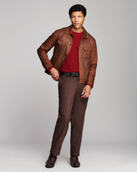 A man modeling a brown leather jacket over a dark red crewneck shirt, paired with brown trousers and dark brown leather shoes, against a light gray studio background.