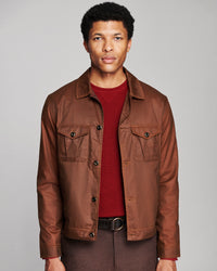 A man wearing a brown waxed cotton utility jacket over a dark red knit sweater, paired with brown trousers and a black belt, photographed against a neutral gray studio background.