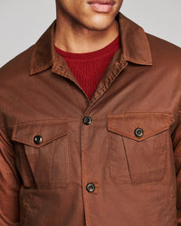 A close-up of a man wearing a brown waxed cotton utility shirt jacket over a red knit crewneck sweater, against a neutral gray background.