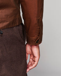 Close-up of a person wearing a brown waxed cotton jacket and dark brown trousers, showing the sleeve cuff with a button tab and the hand.