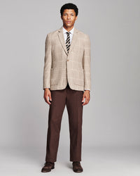 A man modeling a beige and brown plaid blazer with dark brown trousers, a white dress shirt, a striped tie, and brown dress shoes, standing against a light gray studio background.