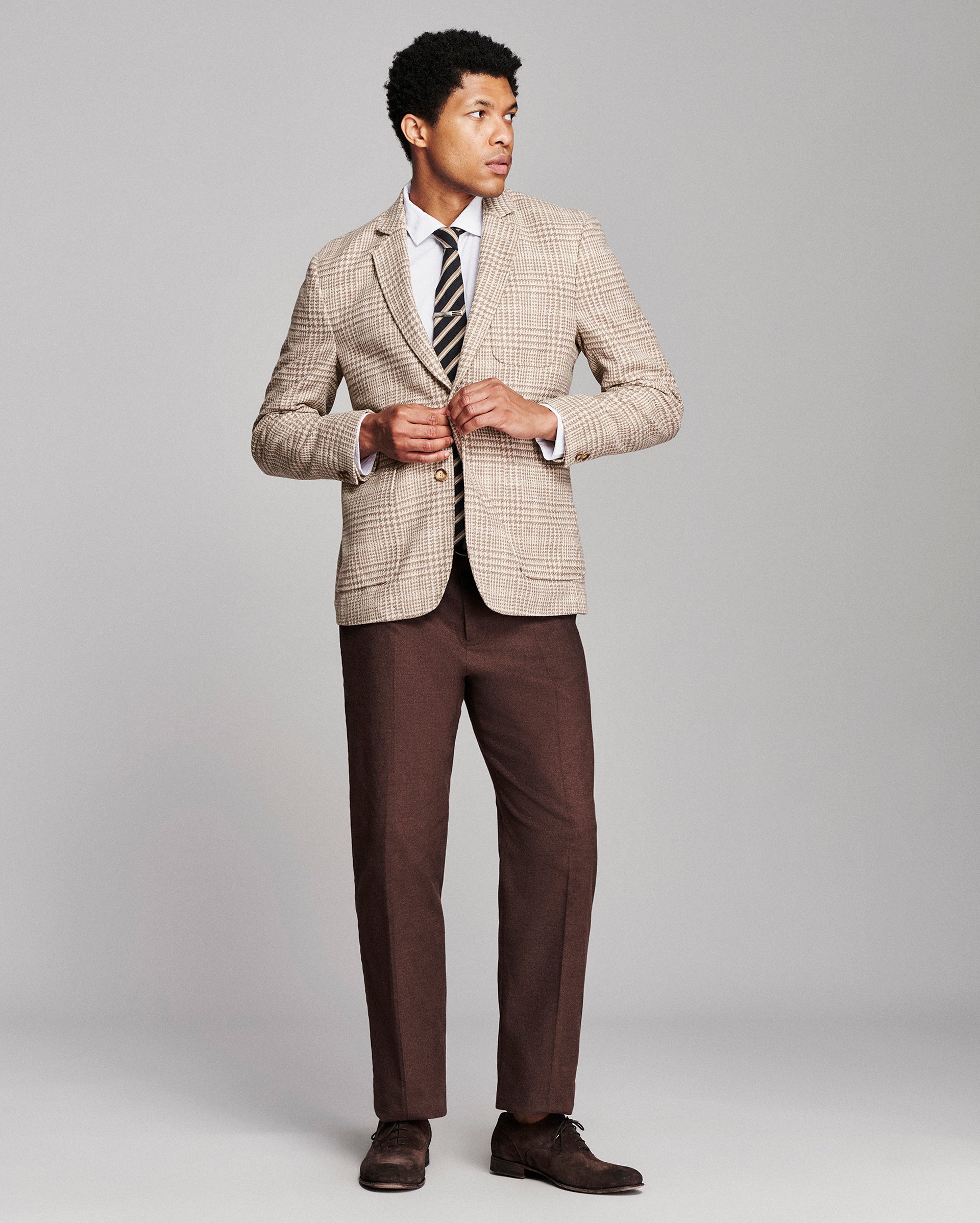 A man modeling a beige and brown checkered blazer, brown trousers, a white dress shirt, a striped tie, and brown dress shoes against a solid gray studio background.