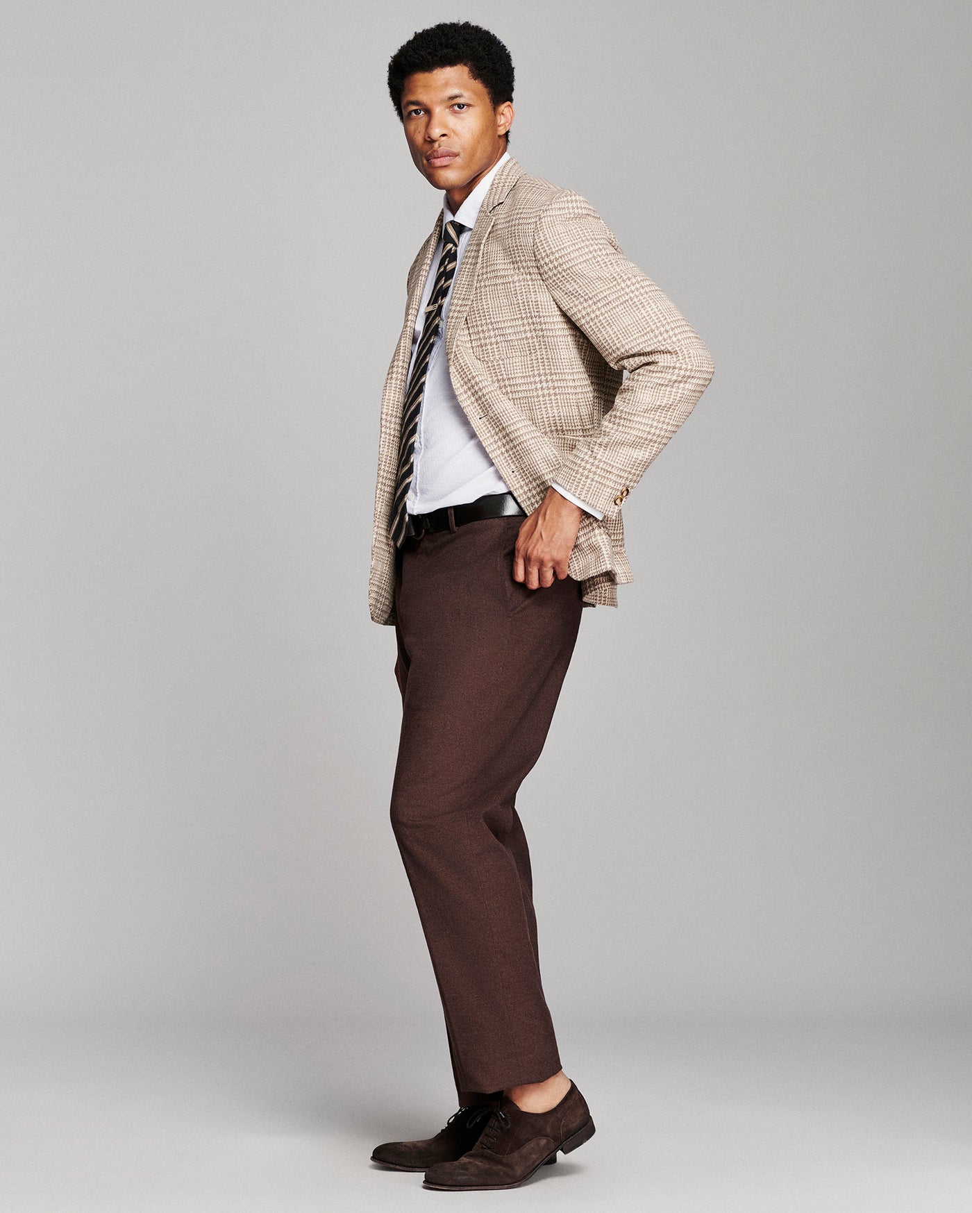 A man modeling a beige and brown checkered blazer, white dress shirt, striped tie, dark brown trousers, and dark brown suede loafers against a light gray studio background.
