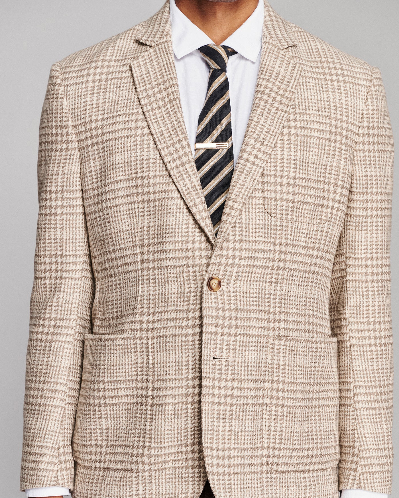 A men's single-breasted suit jacket in a beige and brown houndstooth pattern, styled with a white dress shirt and a dark striped tie, shown on a model against a neutral gray background.