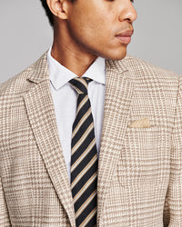 A close-up of a man wearing a beige and brown houndstooth patterned blazer, a white dress shirt, and a dark striped tie against a neutral gray background.
