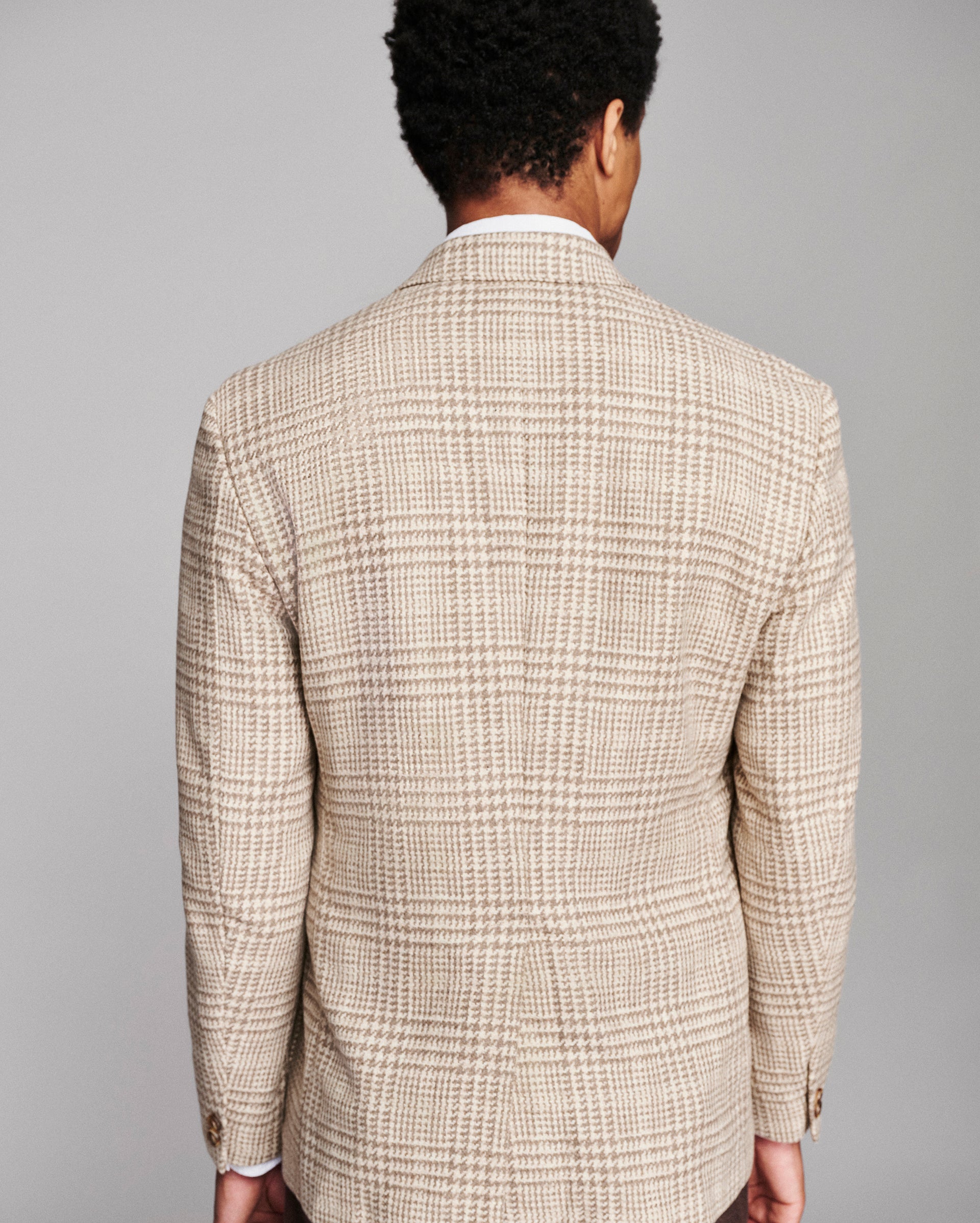 A man viewed from behind wearing a beige and brown plaid blazer over a white collared shirt, set against a solid gray studio background.