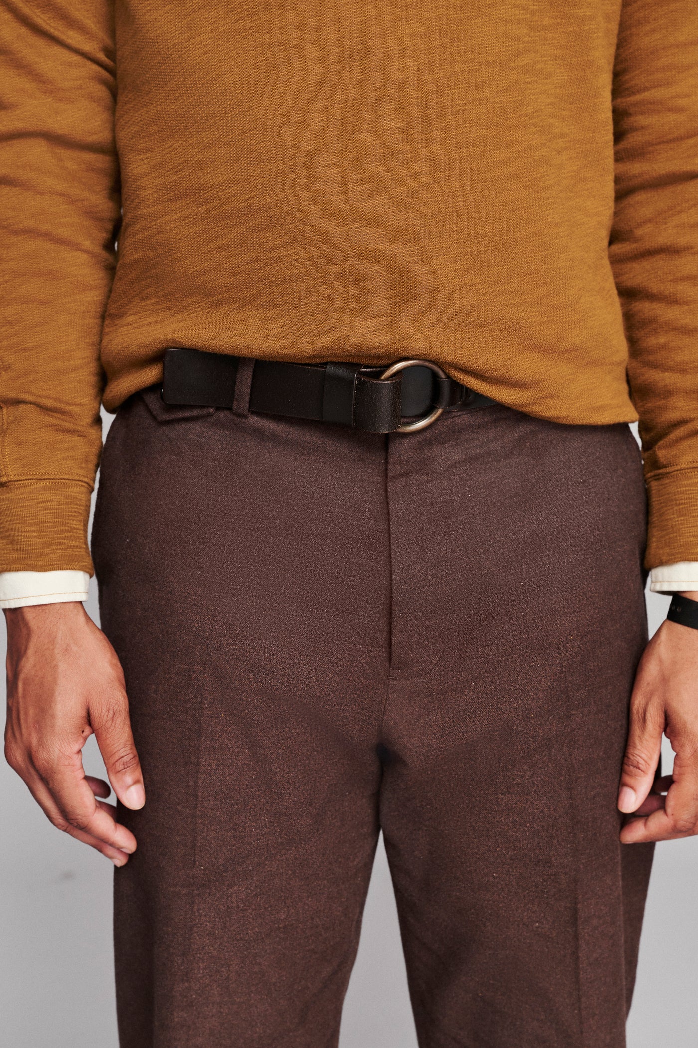 A close-up of a person wearing textured brown trousers, a black leather belt with a silver-tone oval buckle, and a mustard yellow knit sweater.