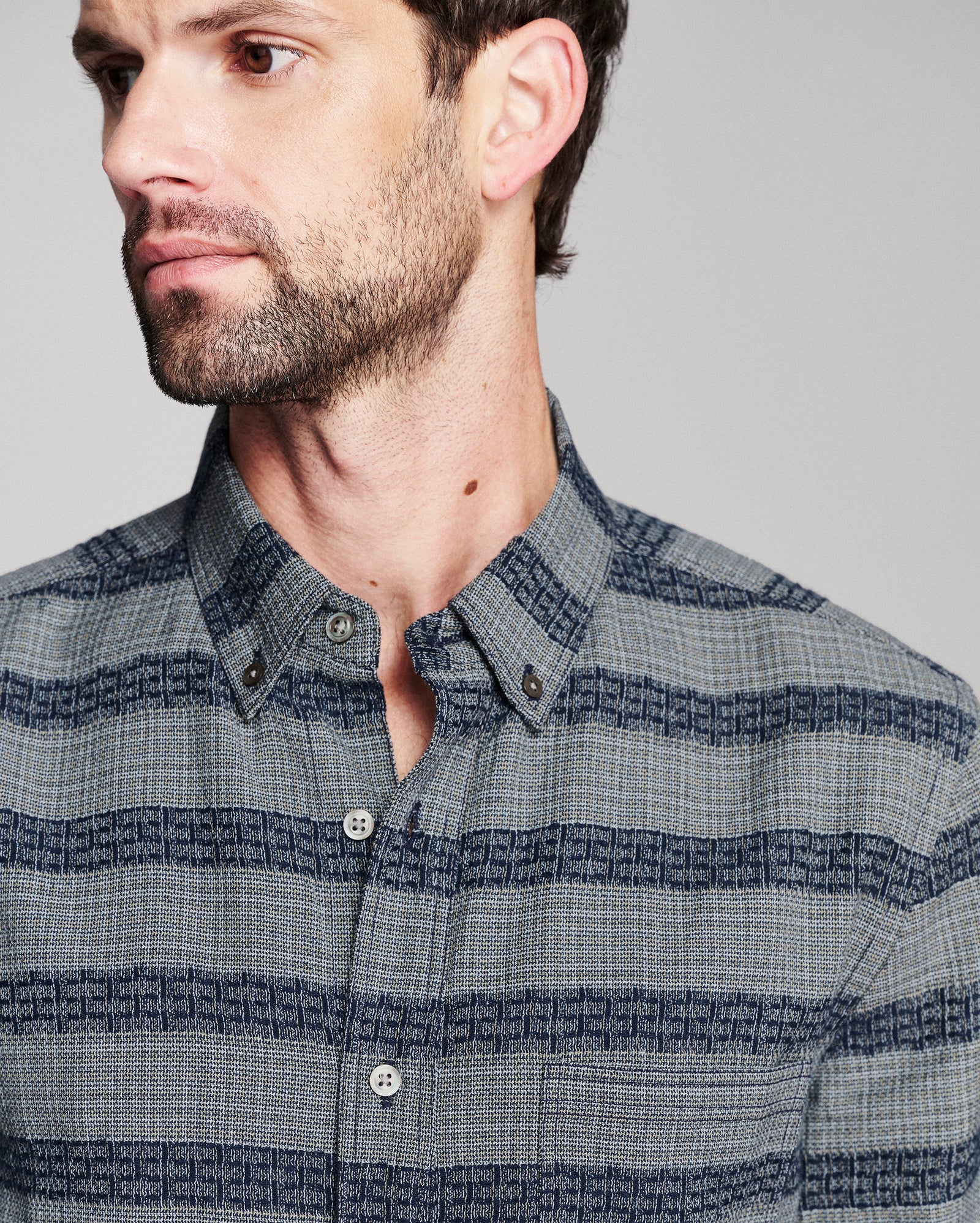 A close-up of a man wearing a blue and gray striped button-down shirt with a subtle geometric pattern.