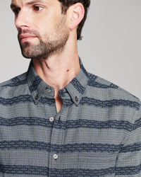 A close-up of a man wearing a blue and gray striped button-down shirt with a subtle geometric pattern.