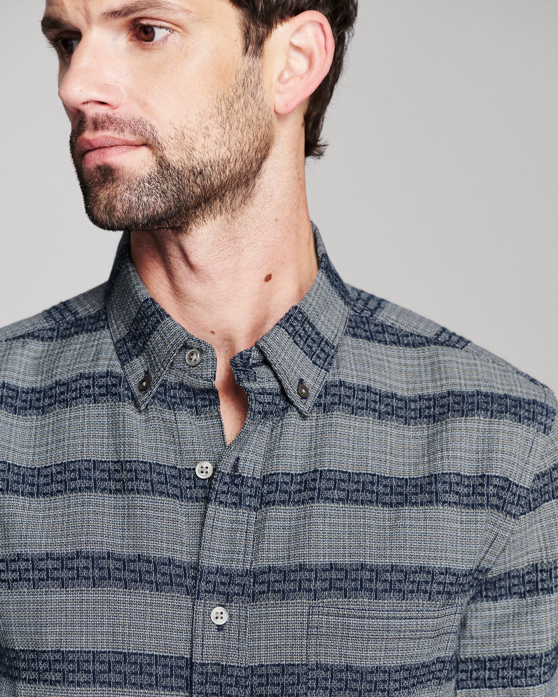 A close-up of a man wearing a blue and gray striped button-down shirt with a subtle geometric pattern.