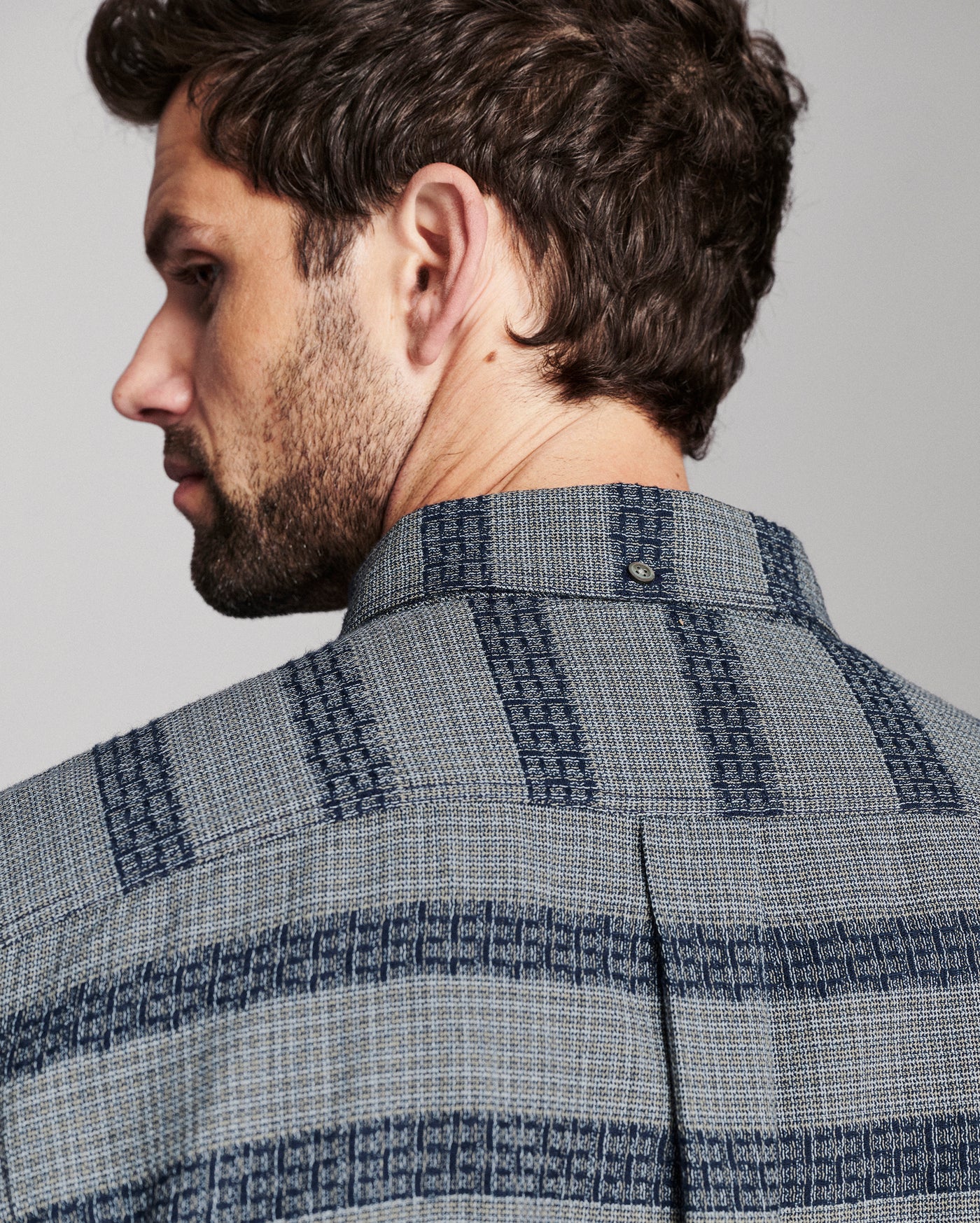 Close-up side profile of a man wearing a textured blue and gray patterned button-down shirt, showing the collar and fabric weave against a neutral gray background.