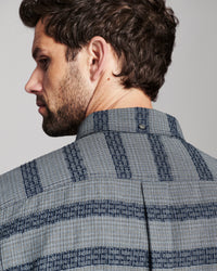 Close-up side profile of a man wearing a textured blue and gray patterned button-down shirt, showing the collar and fabric weave against a neutral gray background.