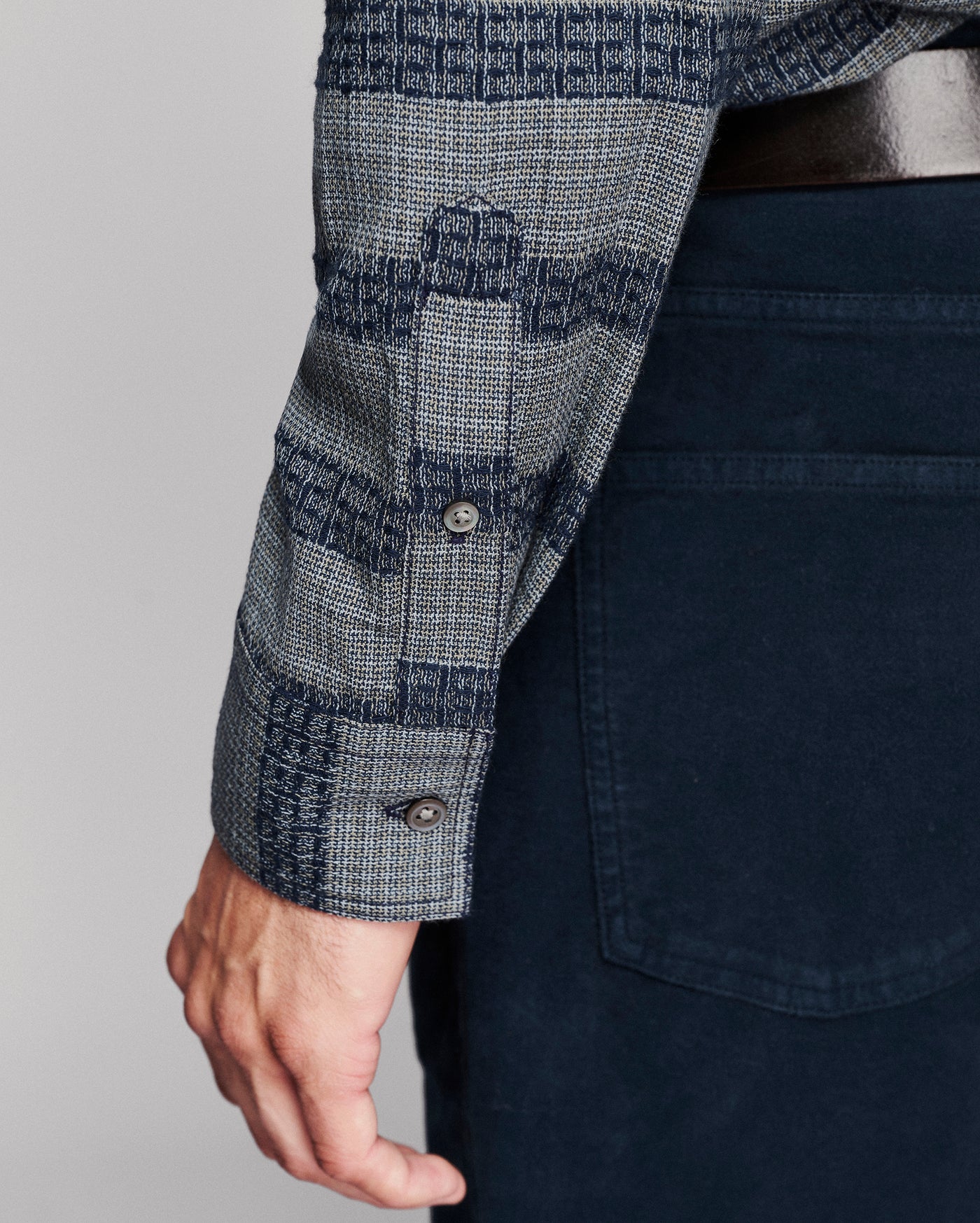 Close-up of a person wearing a patterned long-sleeve button-up shirt with dark navy pants and a dark belt, shown against a light gray studio background.