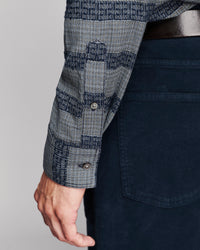 Close-up of a person wearing a patterned long-sleeve button-up shirt with dark navy pants and a dark belt, shown against a light gray studio background.