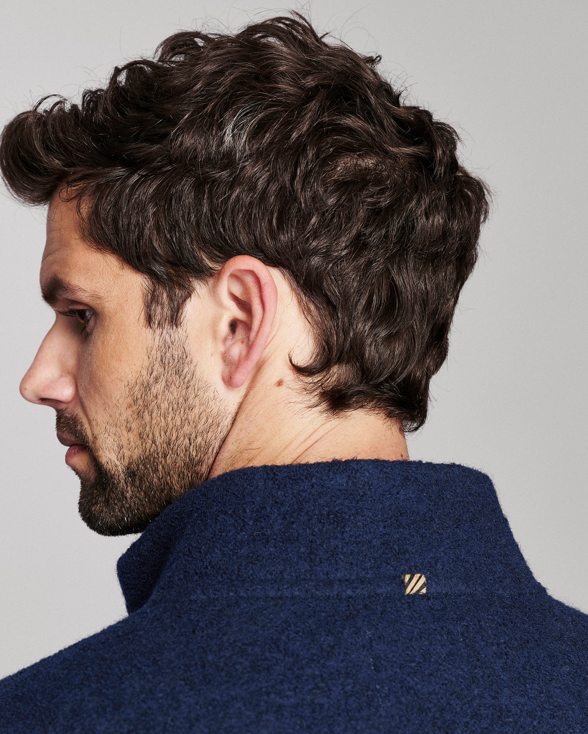 Close-up side profile of a man with dark curly hair wearing a navy blue textured knit sweater, showing a small gold embroidered logo on the collar against a light gray background.