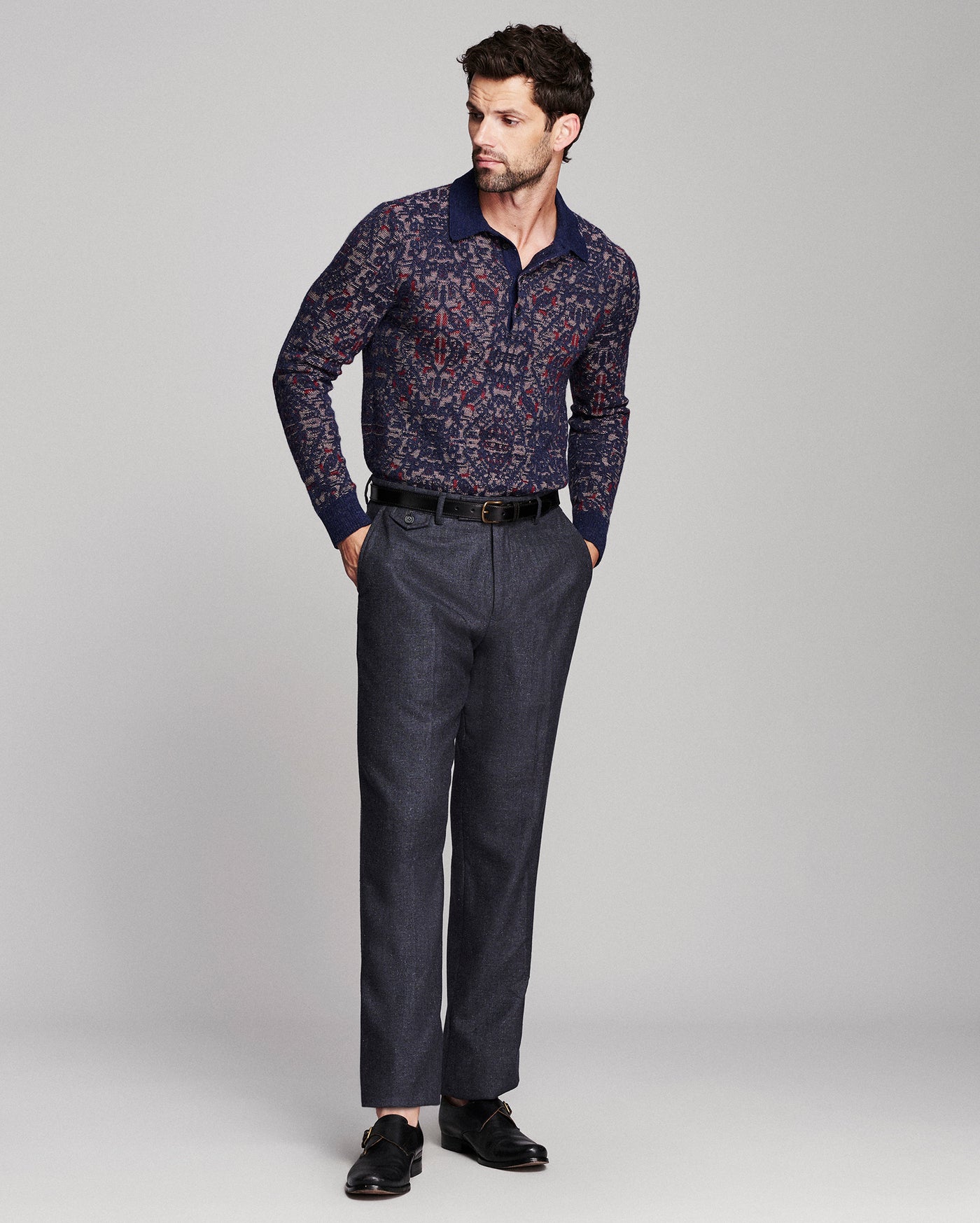 A man modeling a long-sleeve collared shirt with a navy and red abstract pattern, paired with dark gray trousers and black loafers, against a light gray studio background.