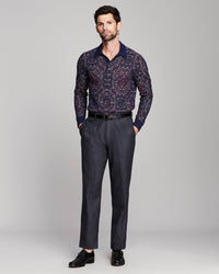 A man modeling a long-sleeve button-up shirt with a navy blue and red geometric pattern, paired with dark gray trousers and black dress shoes, against a light gray studio background.