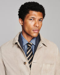 A man wearing a light beige overshirt over a blue shirt and a striped tie, photographed against a plain light gray studio background.