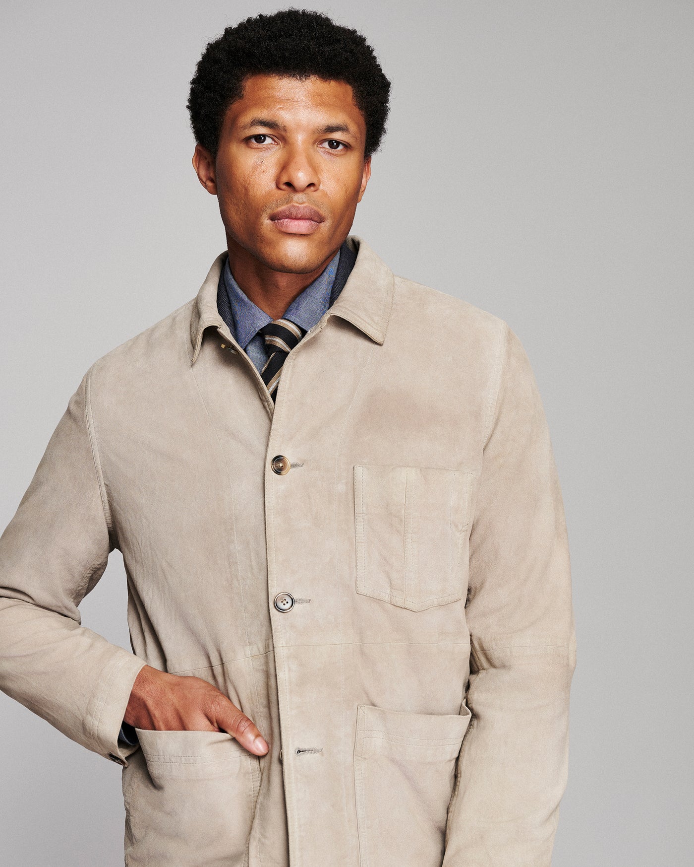 A man wearing a light beige corduroy utility jacket over a blue collared shirt and striped tie, photographed against a solid gray studio background.