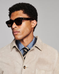 A man wearing black square sunglasses, a beige overshirt, a blue shirt, and a striped tie, photographed against a solid gray background.