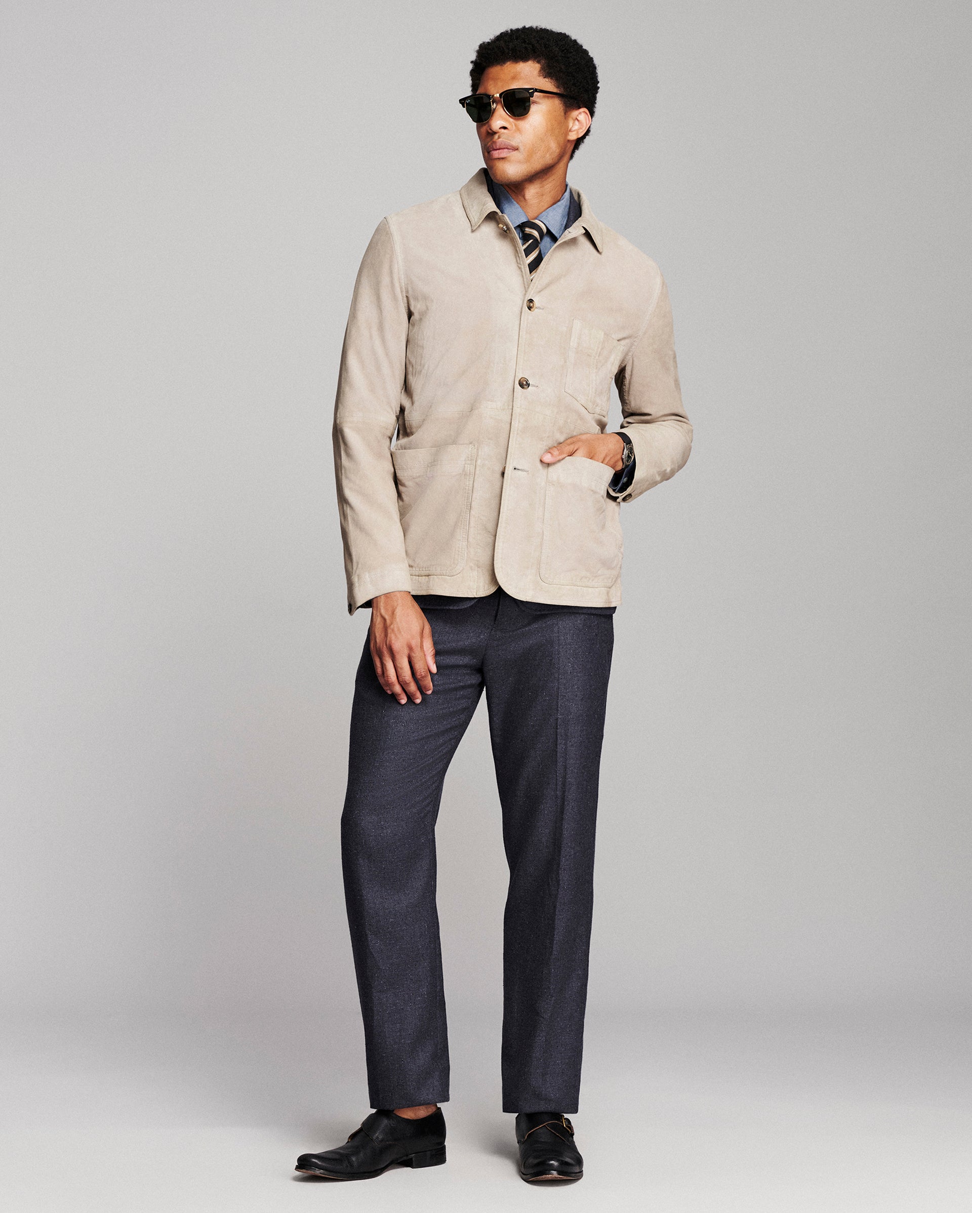 A man modeling a light beige corduroy jacket over a blue patterned shirt, paired with navy trousers and black dress shoes, against a light gray studio background.