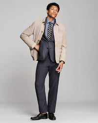 A man modeling a beige overshirt over a navy blue suit, including a blue dress shirt, striped tie, and navy trousers, paired with black leather dress shoes, against a light gray studio background.