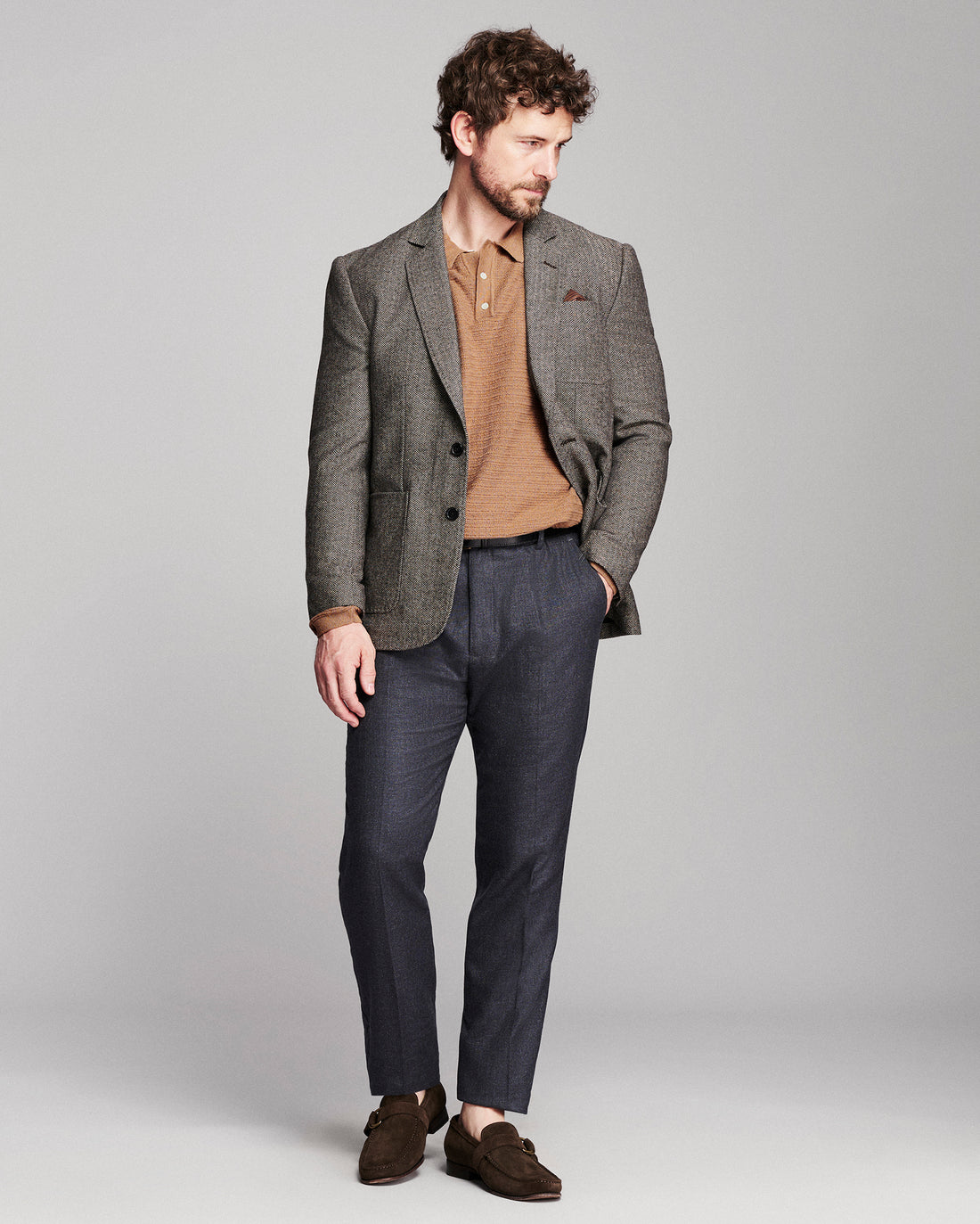 A man modeling a brown textured blazer over a tan knit polo shirt, paired with navy trousers and brown suede loafers, against a solid light gray studio background.