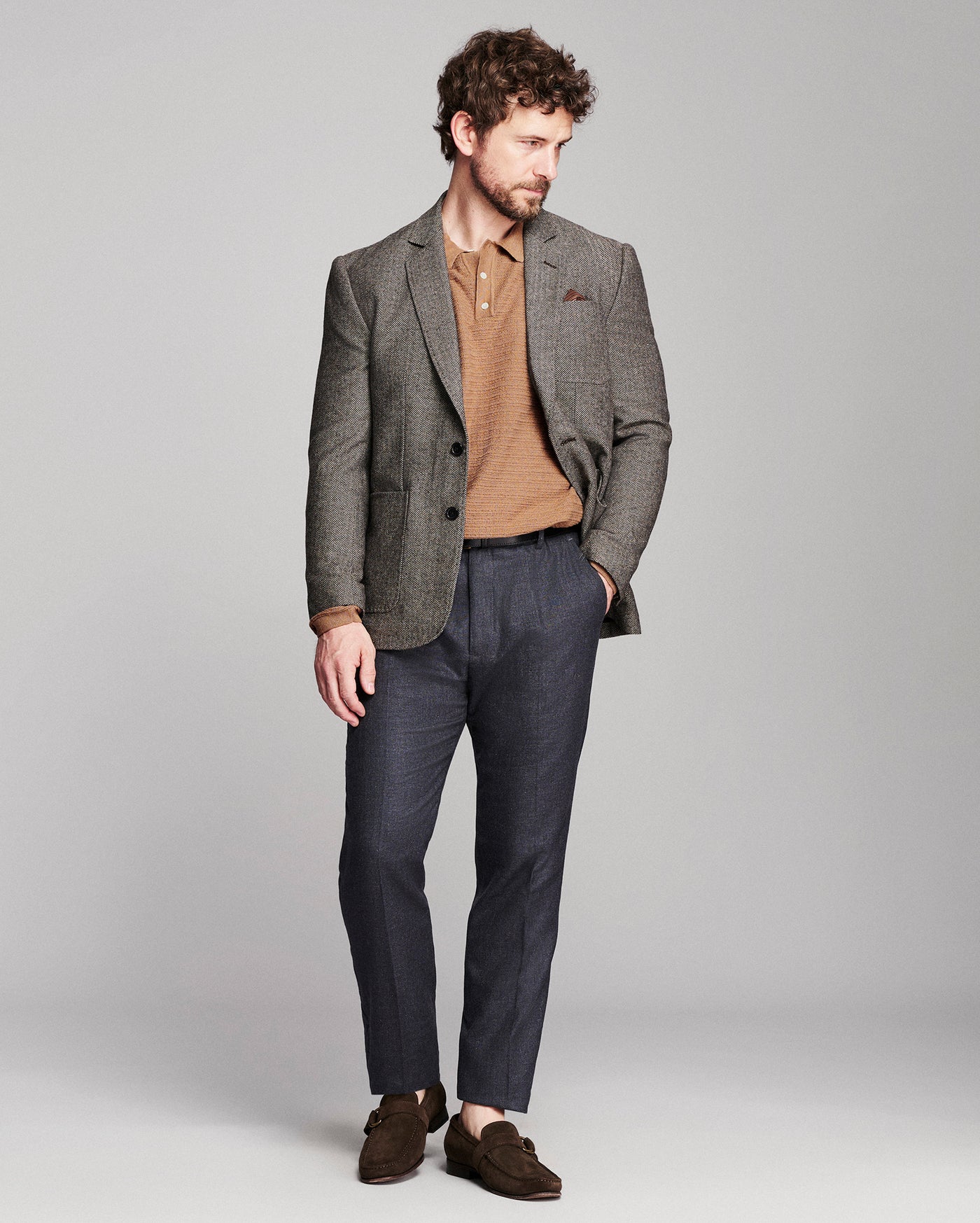 A man modeling a brown textured blazer over a tan knit polo shirt, paired with navy trousers and brown suede loafers, against a solid light gray studio background.