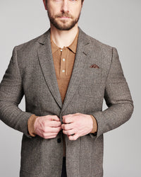 A man wearing a brown and gray textured tweed blazer over a brown polo shirt, shown from the chest up against a neutral gray background.