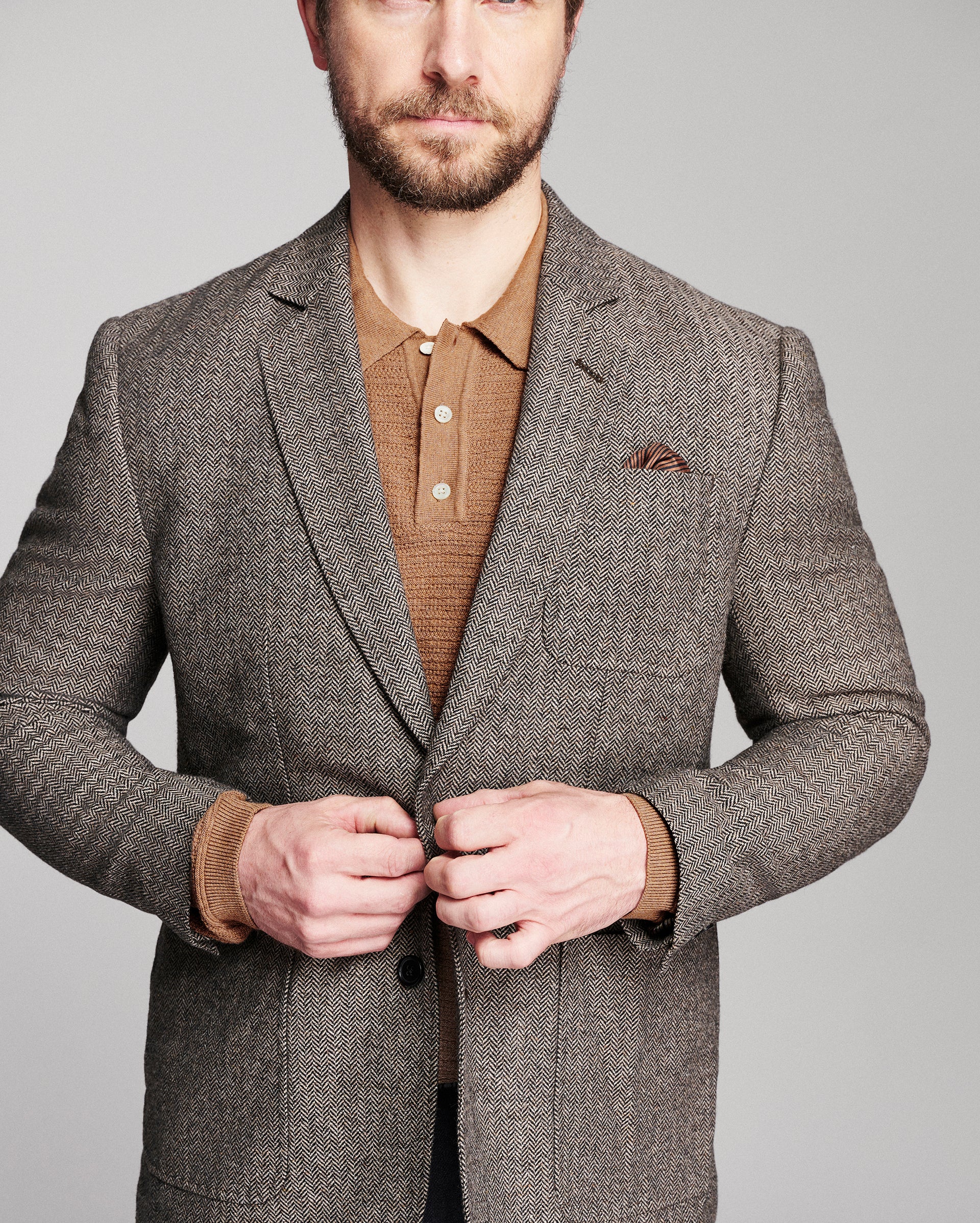 A man wearing a brown and gray textured tweed blazer over a brown polo shirt, shown from the chest up against a neutral gray background.