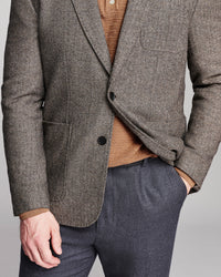 A close-up of a man wearing a textured gray-brown herringbone blazer over a brown knit sweater and dark gray trousers, shown from the chest down against a neutral studio background.