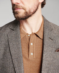 Close-up of a man wearing a brown and gray herringbone patterned blazer over a brown knit polo shirt, against a neutral gray background.