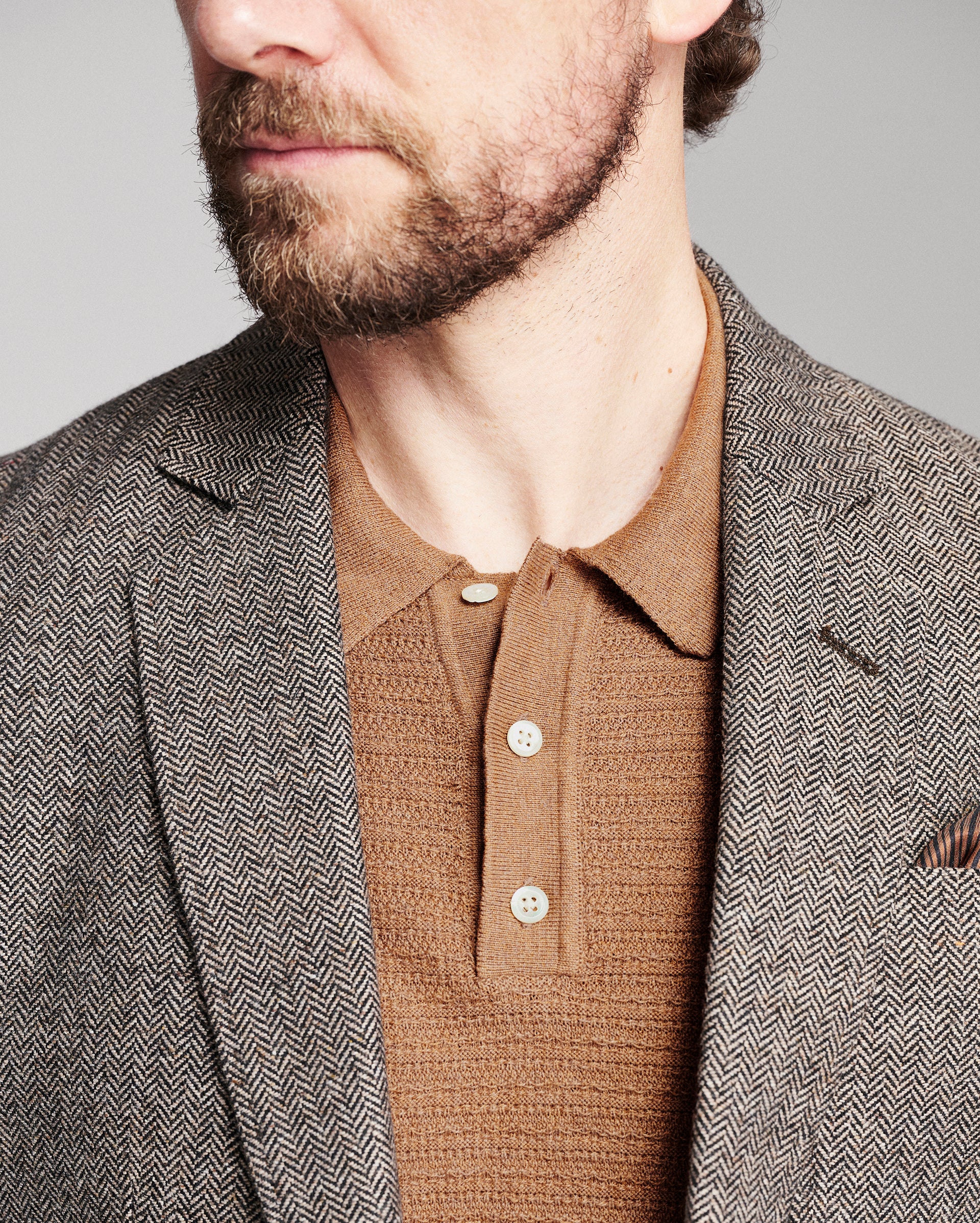 Close-up of a man wearing a brown and gray herringbone patterned blazer over a brown knit polo shirt, against a neutral gray background.