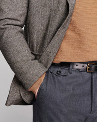 Close-up of a person wearing a gray and brown herringbone patterned blazer over a tan knit sweater, paired with dark gray trousers and a black belt.