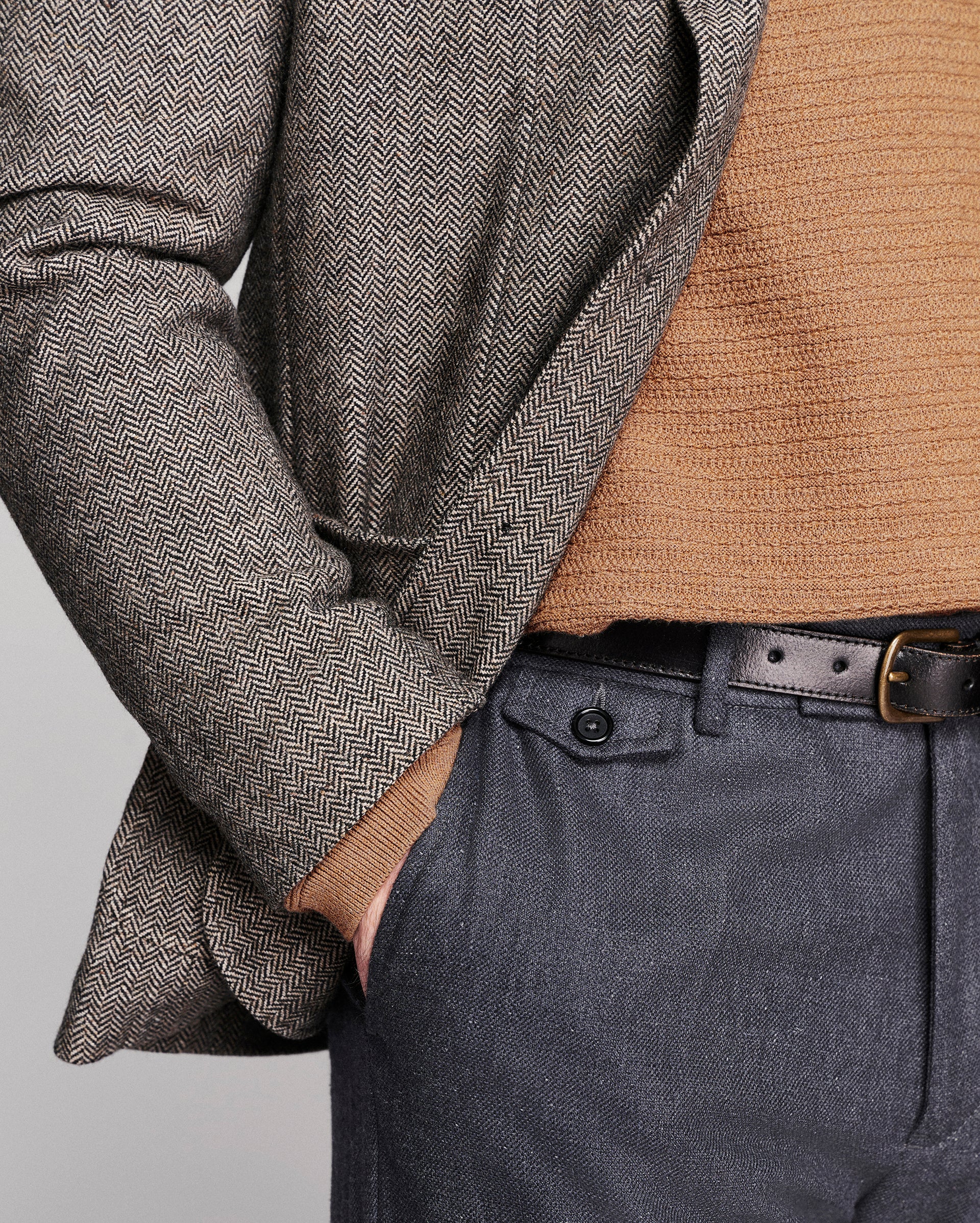 Close-up of a person wearing a gray and brown herringbone patterned blazer over a tan knit sweater, paired with dark gray trousers and a black belt.