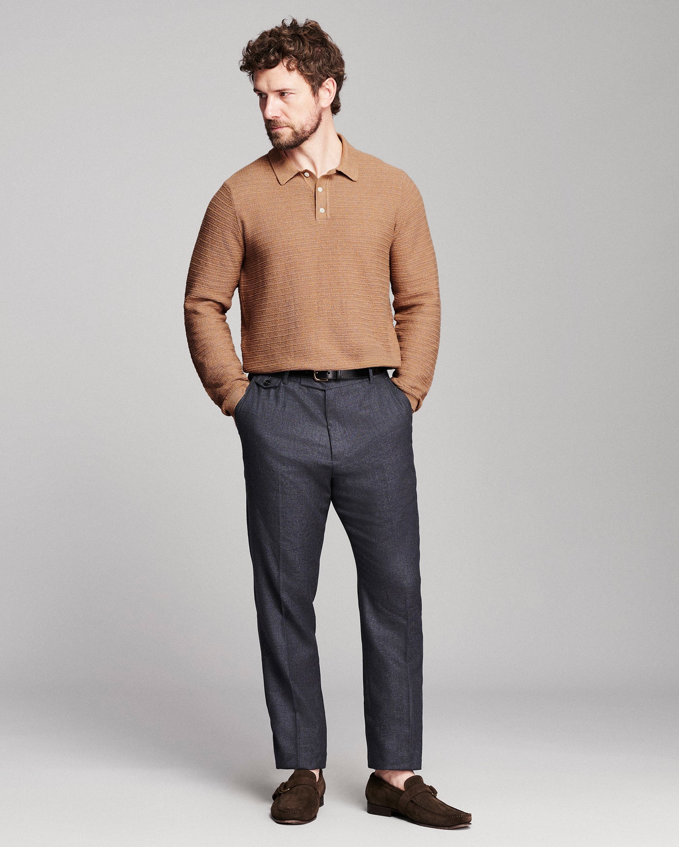 A man modeling a tan textured knit polo sweater, dark gray trousers, and brown suede loafers against a light gray studio background.