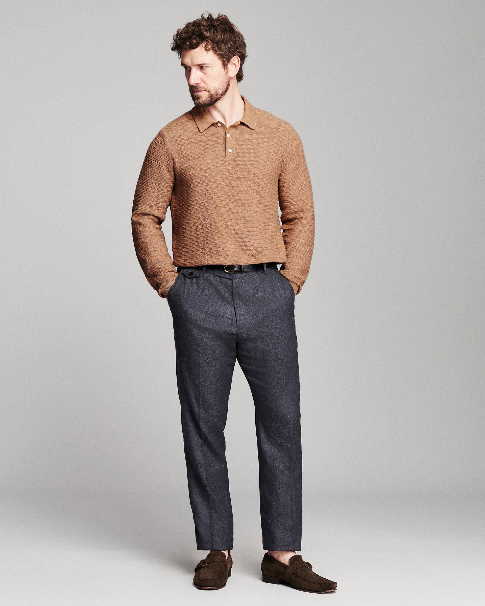 A man modeling a tan textured knit polo sweater, dark gray trousers, and brown suede loafers against a light gray studio background.