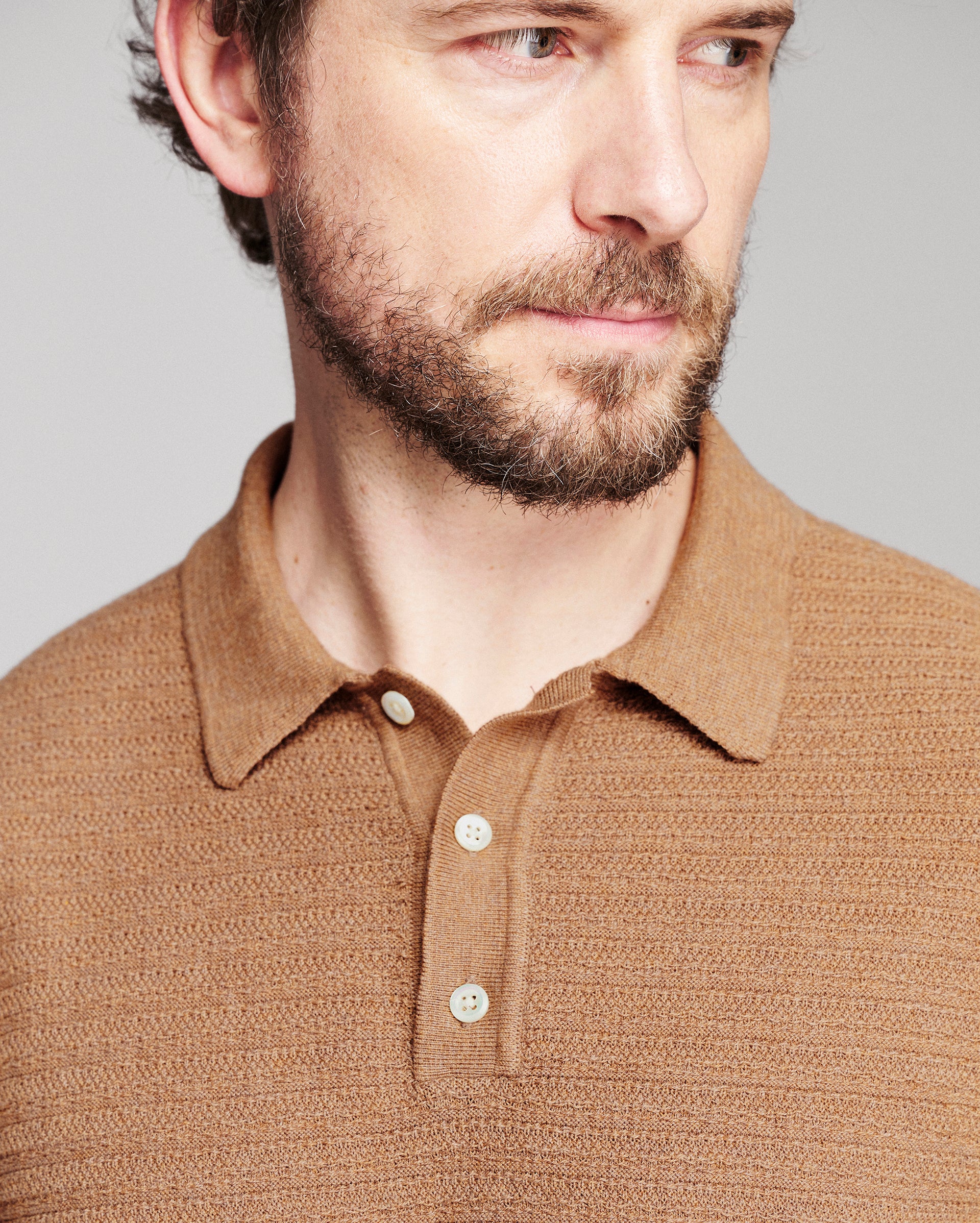 Close-up of a man wearing a textured knit polo shirt in a warm camel brown color, featuring a classic collar and three white buttons.